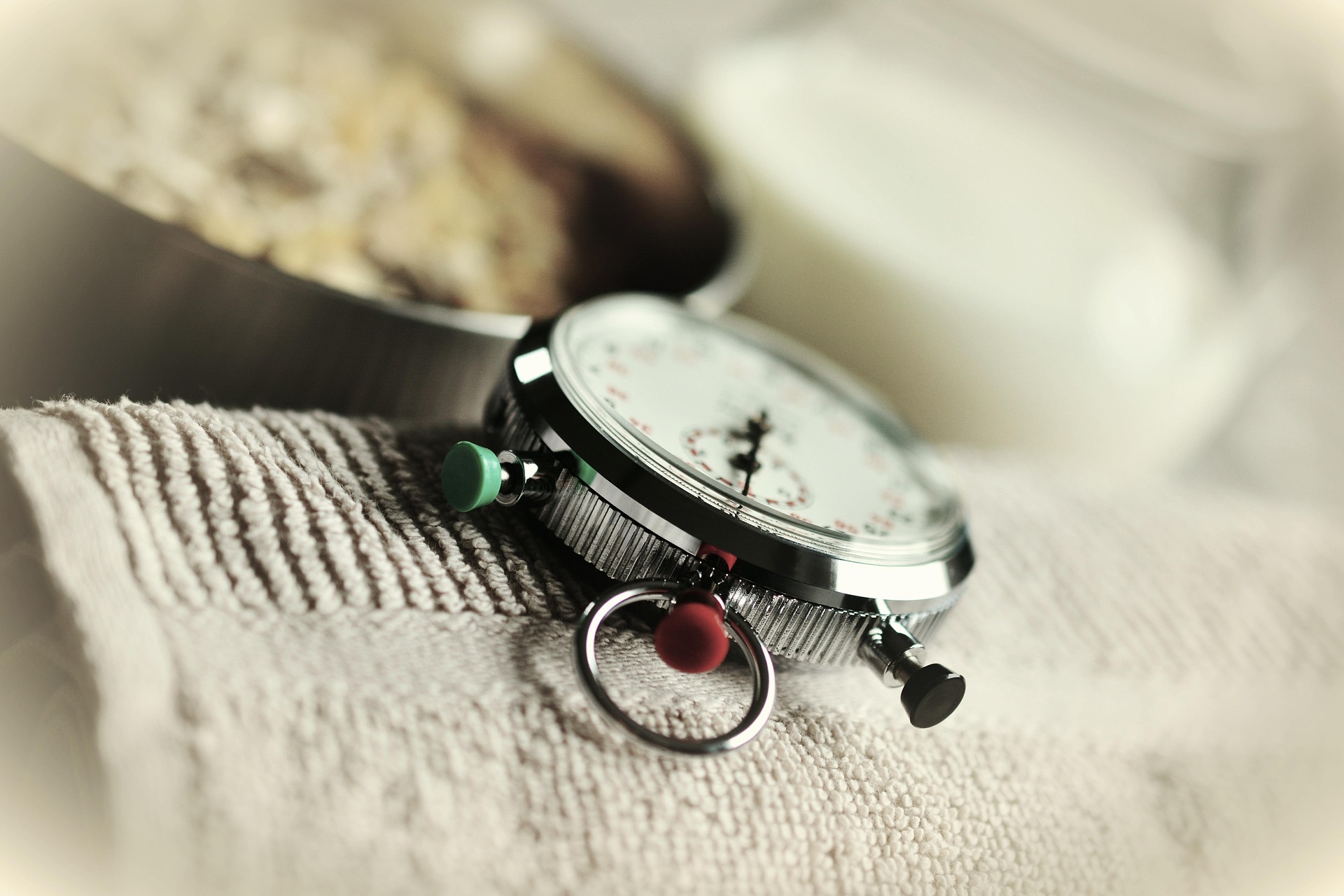 A silver stopwatch with a green and a red push button and a metal ring, resting on a pale towel, with a bowl of muesli and a glass of milk blurred behind it